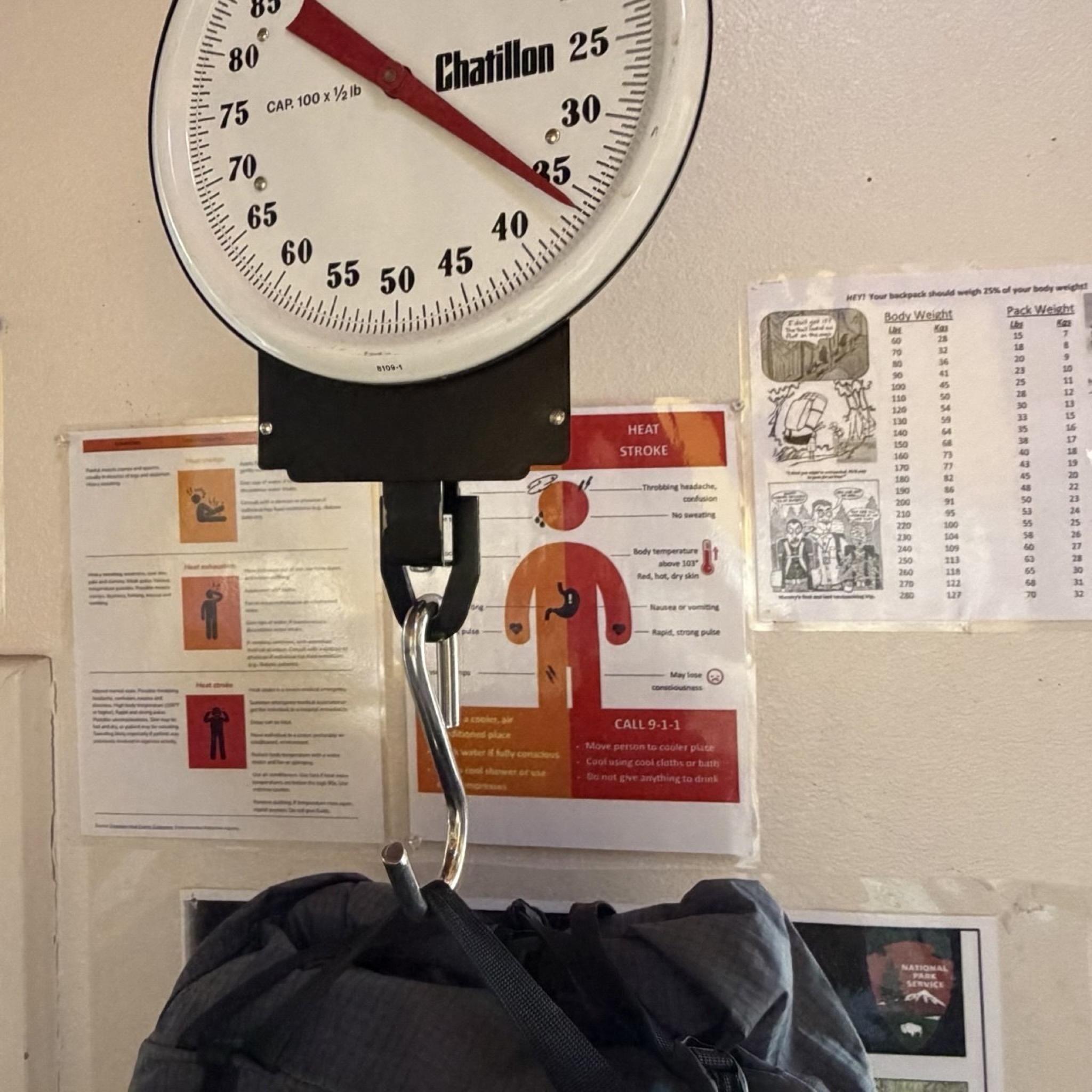 A hanging scale weighing a backpack next to heat-stroke and pack-weight warning posters at the backcountry information center