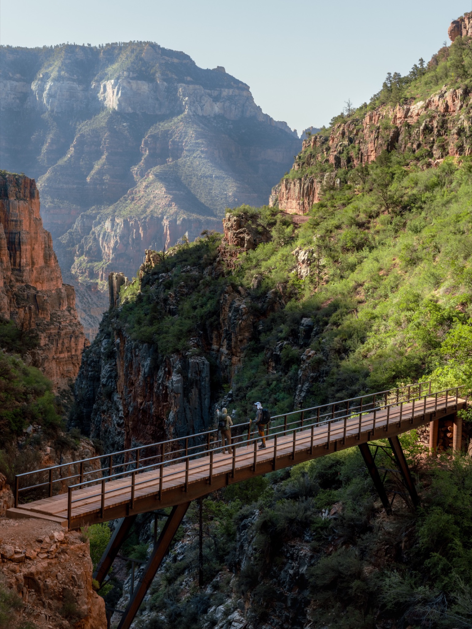 Two hikers crossing a footbridge deep in the Grand Canyon beneath towering cliffs