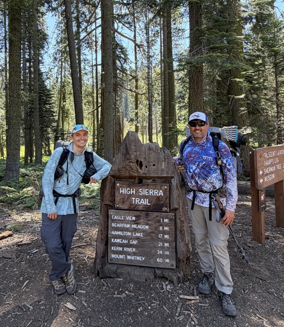 Two backpackers standing beside the wooden High Sierra Trail trailhead sign at Crescent Meadow listing mileages to Bearpaw Meadow, Hamilton Lake, Kaweah Gap and Mount Whitney
