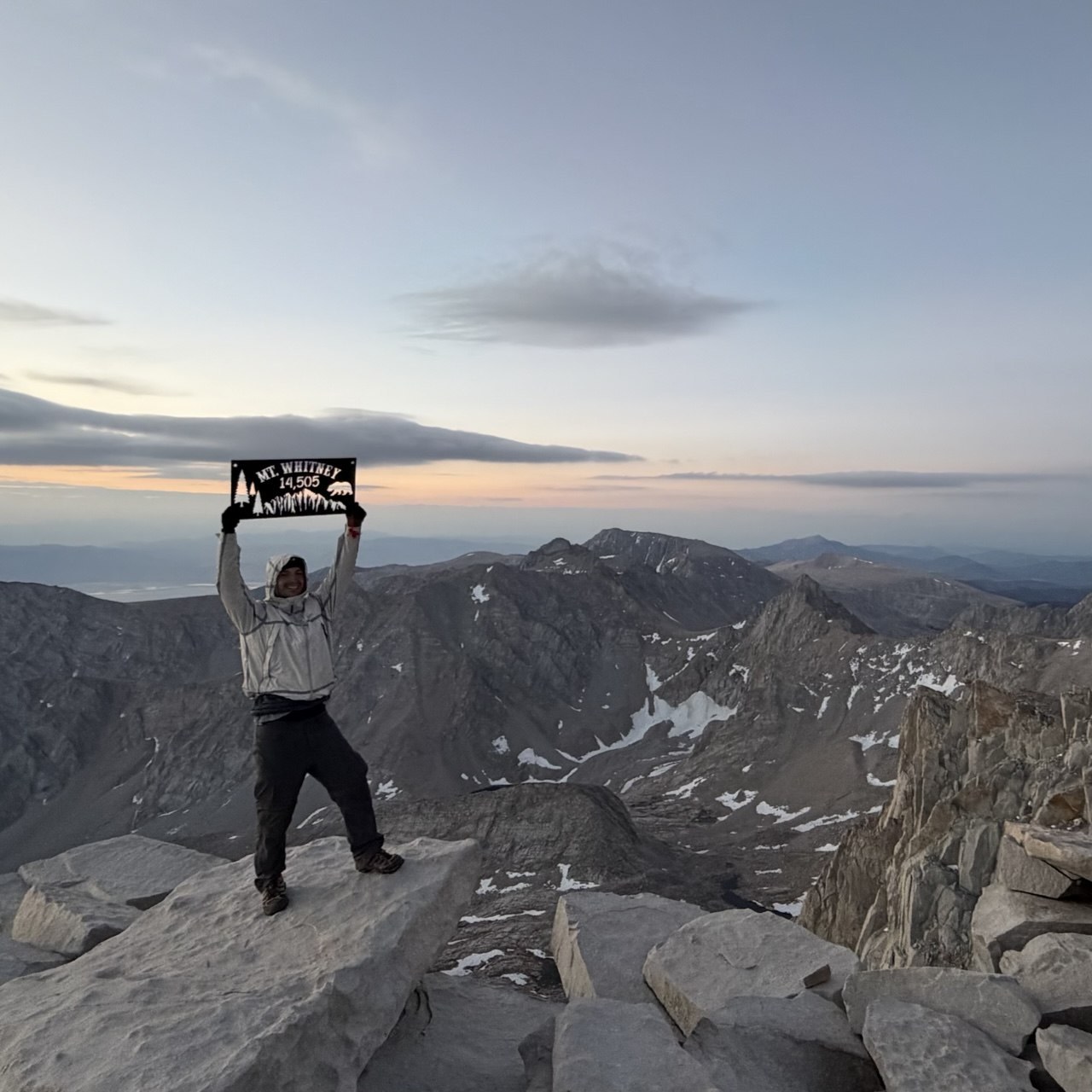 A hiker holding a Mount Whitney 14,505 summit sign at sunrise on the summit plateau