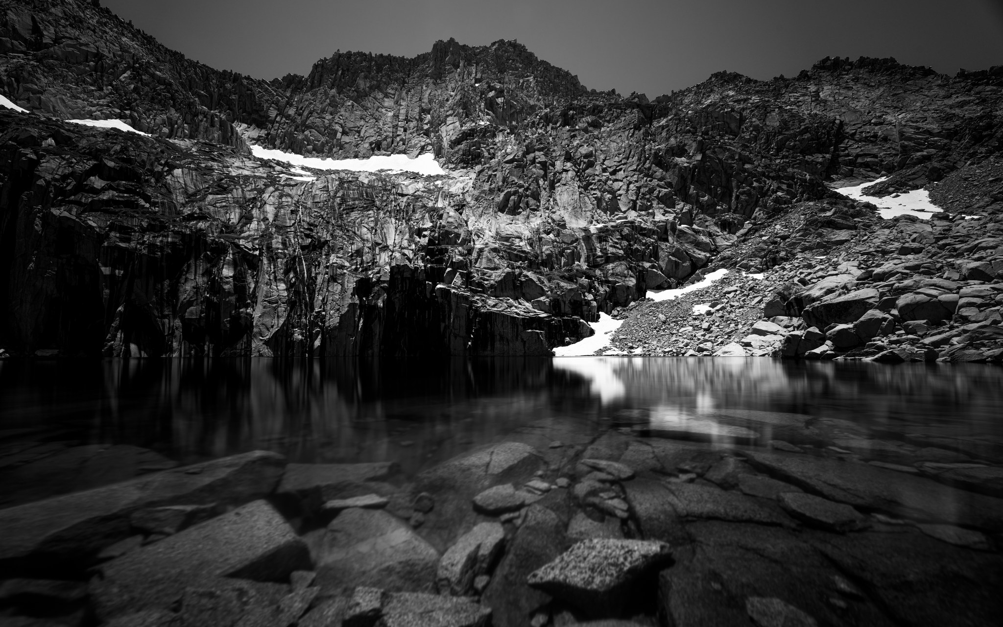 Black and white long exposure of Precipice Lake below the dark cliffs of Eagle Scout Peak on the High Sierra Trail, snow patches above still water