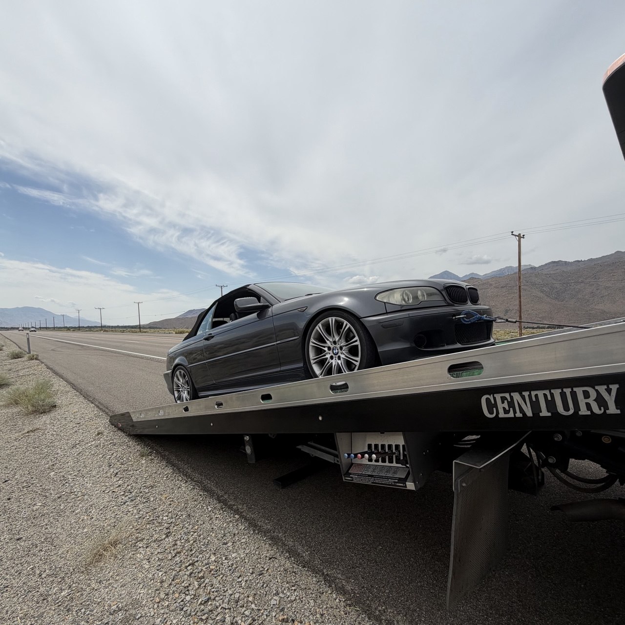 A grey BMW coupe strapped onto a flatbed tow truck on the shoulder of Highway 395 near Bishop, the Sierra Nevada in the distance