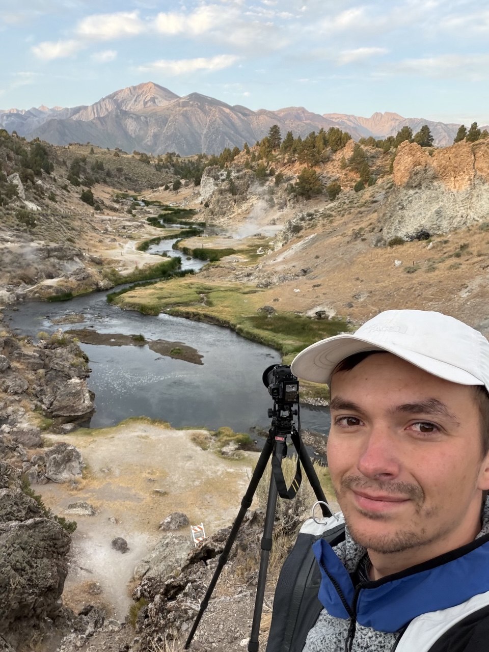 Danil Zanozin with a camera on a tripod above Hot Creek Geological Site at sunrise, steam rising from the creek and alpenglow on the mountains behind Mammoth Lakes