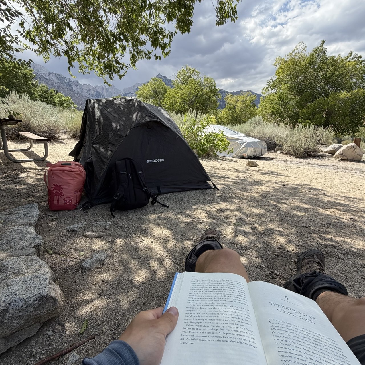 A small tent and backpack at Lone Pine Campground with Mount Whitney and the Sierra crest behind, photographed from a camp chair with an open book