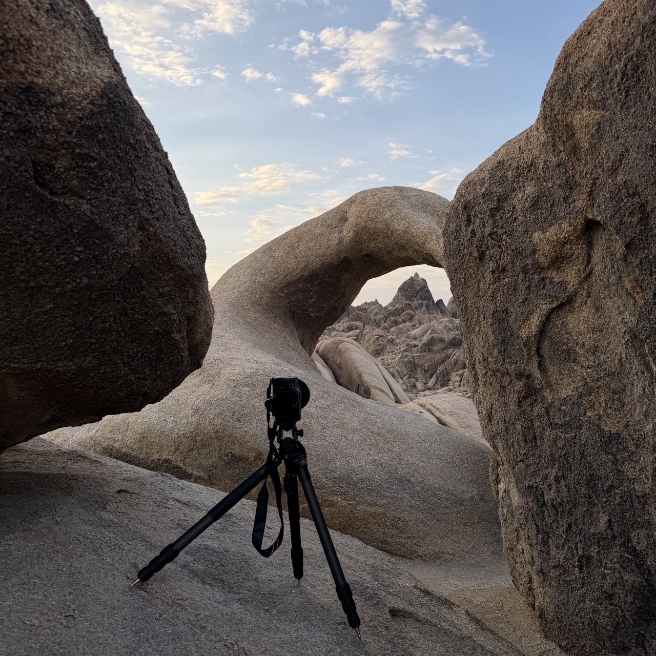 A camera on a tripod set up beneath Mobius Arch in the Alabama Hills at dusk, rock formations visible through the arch