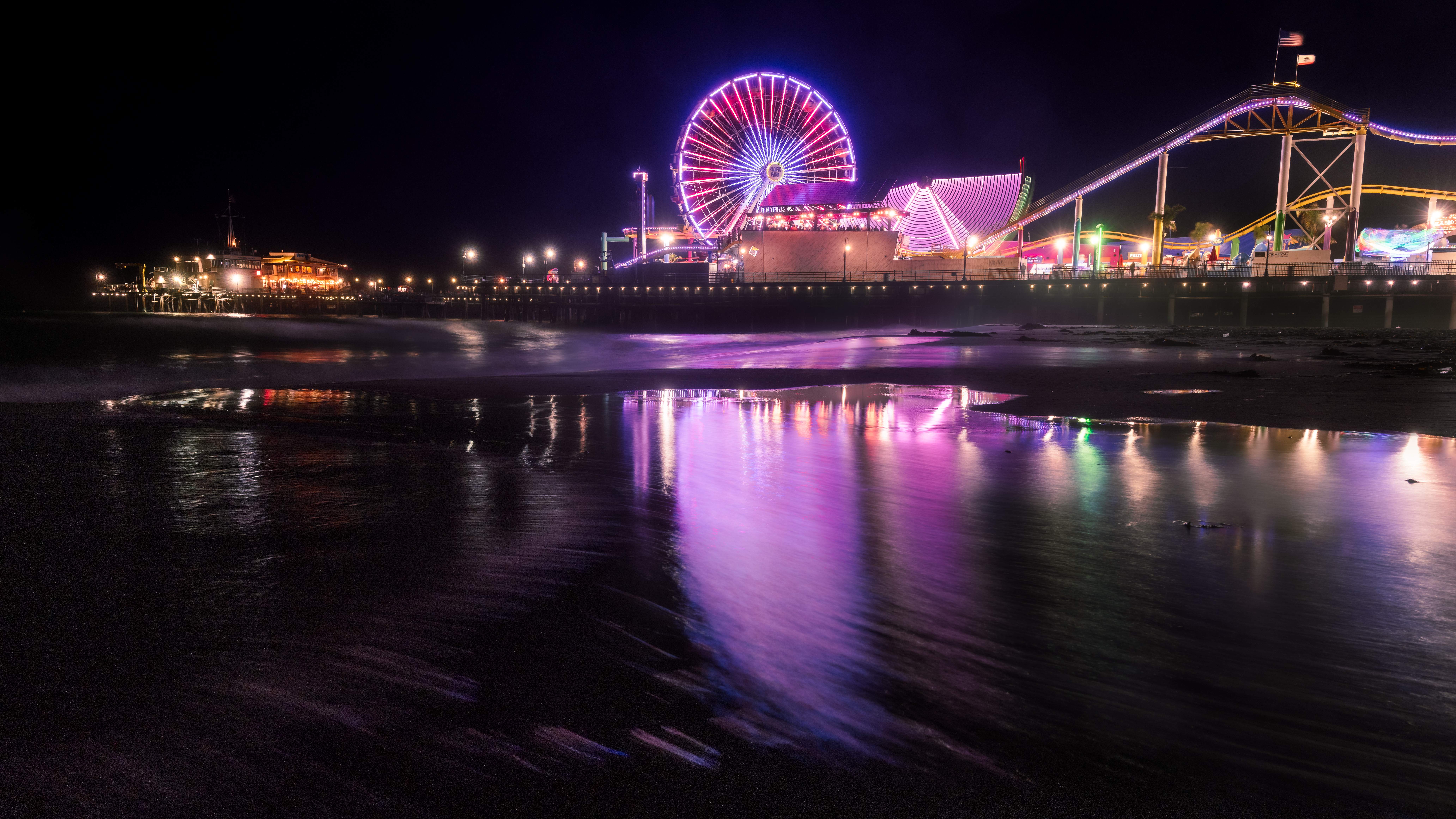 Long-exposure night photo of Santa Monica Pier's lit Ferris wheel and roller coaster reflected on the wet beach.