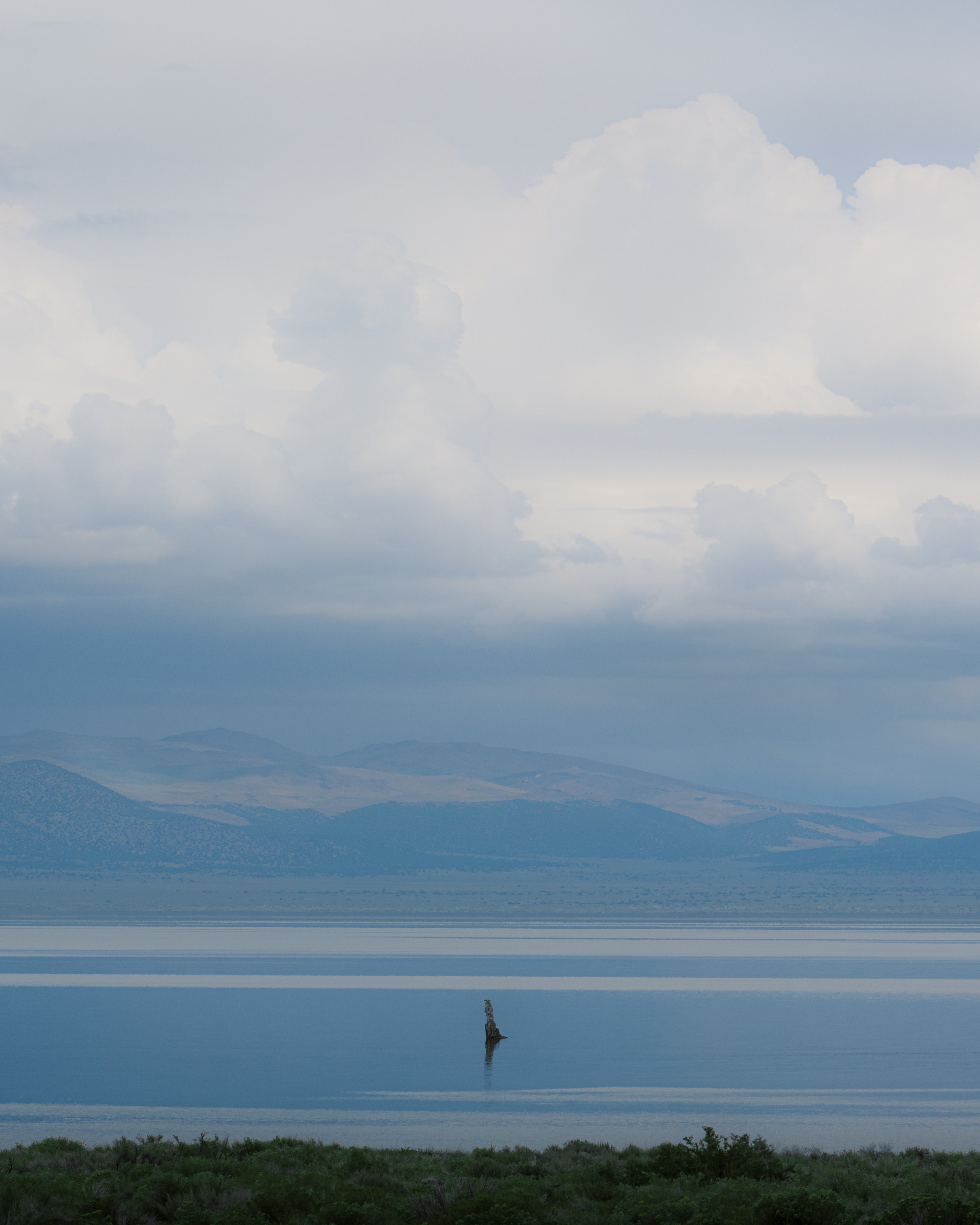 A lone tufa tower in Mono Lake under stacked storm clouds, water, hills and sky in flat bands of blue and grey, California.