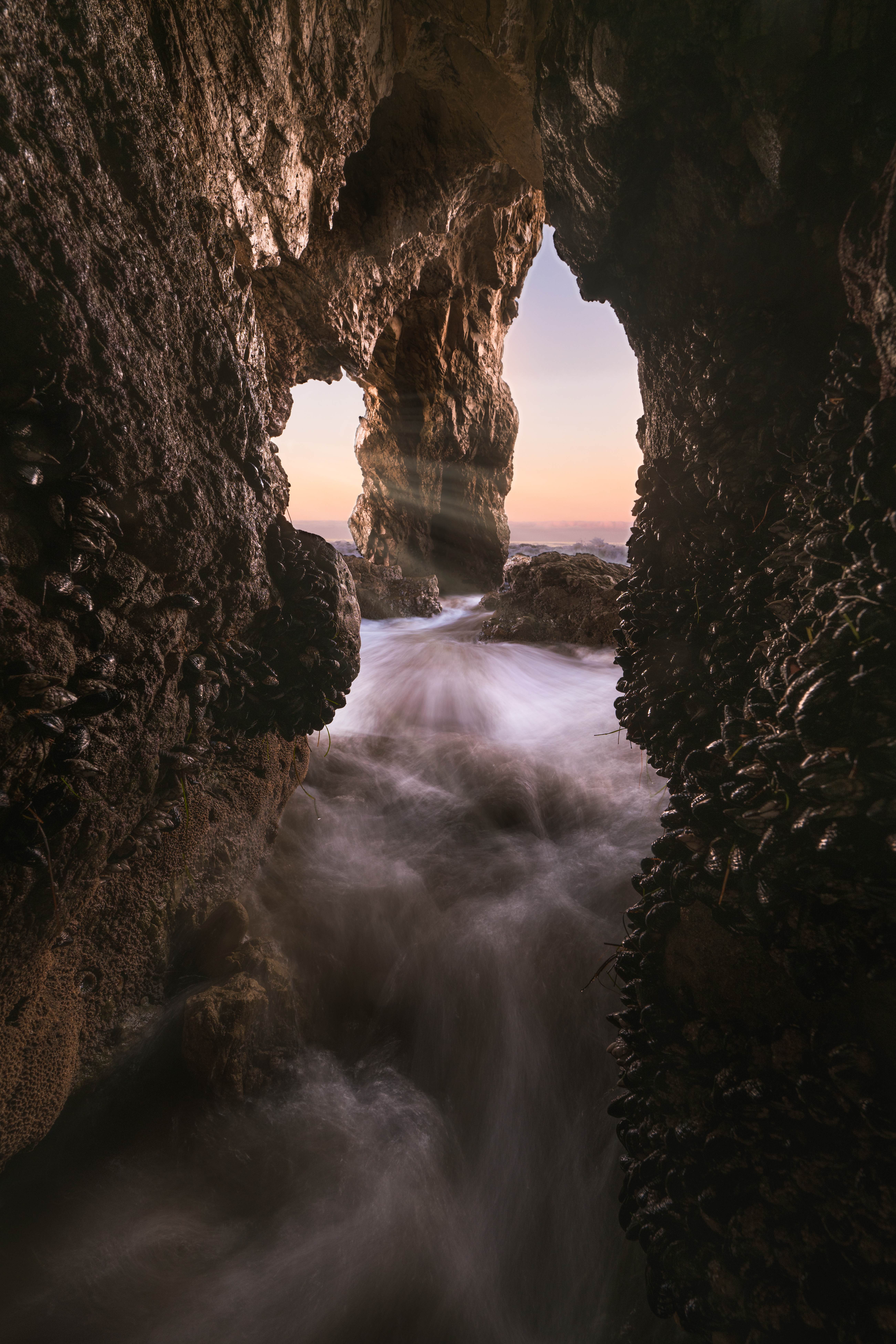 Early-morning light streaming through the sea cave at El Matador Beach, god rays catching in the fog above a low-tide shoreline.